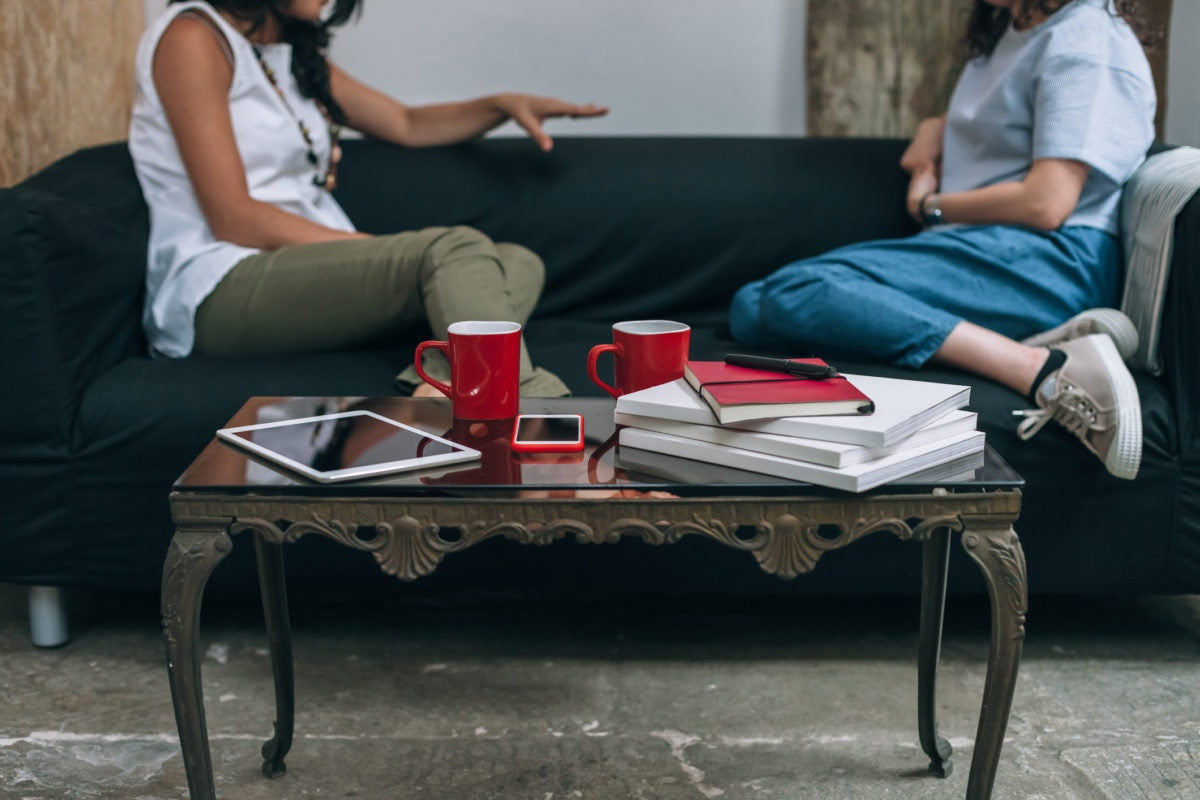 Two girls sitting in a sofa with cups ,tab and books placed on a table